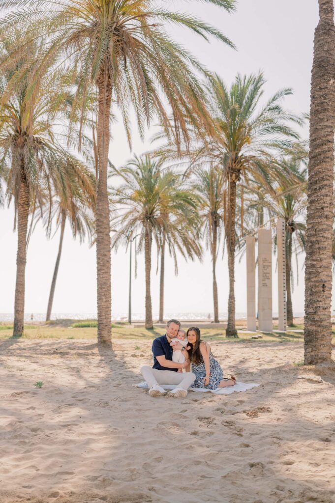 Parents sitting together and lifting their baby into the air beneath the palm trees at Patacona Beach