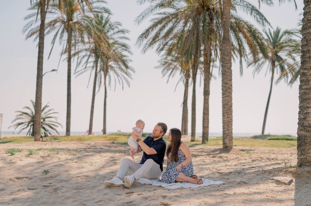 Parents sitting together and lifting their baby into the air beneath the palm trees at Patacona Beach